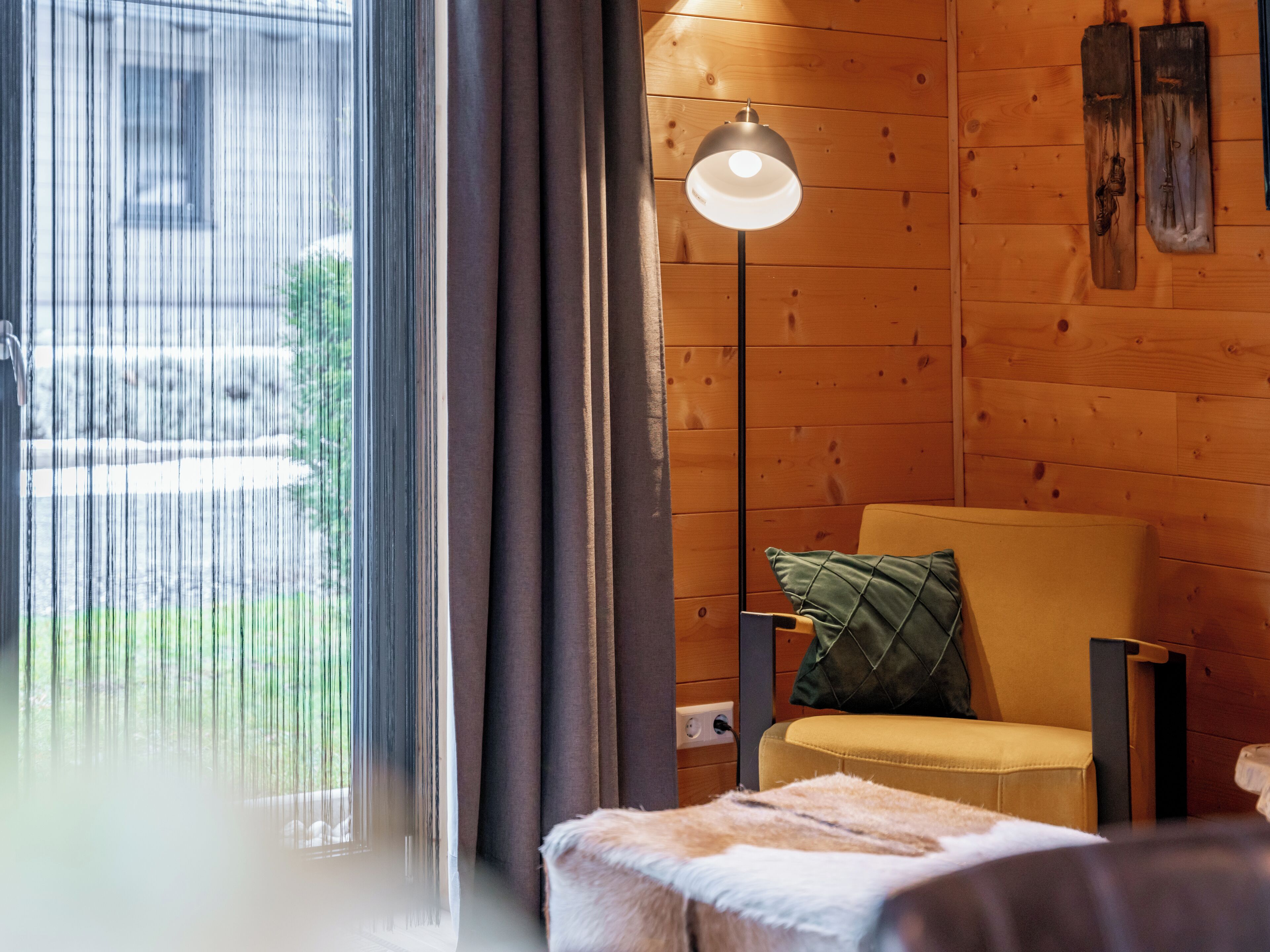 A cozy room with a wooden wall, a floor lamp, a chair with a cushion, and a view of a building outside.
