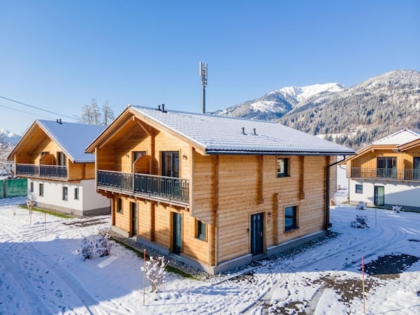 Eine Holzhütte mit einem Balkon, umgeben von verschneitem Gelände und Bergen im Hintergrund.