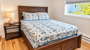 A bedroom with a wooden bed frame, a nightstand with a lamp, and a window with a view of a blue house and greenery. - Modern 2 Bedroom house in ANC (Anchorage)