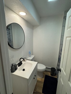 Une petite salle de bain avec un lavabo blanc, un miroir rond, des toilettes et une poubelle noire.