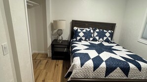 Une chambre avec un plancher en bois, un lit recouvert d’une courtepointe, une table de chevet avec une lampe et une porte donnant sur une autre pièce.