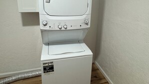 Un appareil compact, blanc, comprenant une machine à laver et un sèche-linge, installé dans un coin d’une pièce dotée d’un plancher en bois et d’un mur blanc.