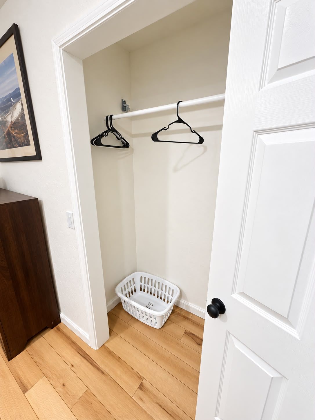 A hallway with a white door, a wooden floor, a white laundry basket, and two black hangers on a white rod.