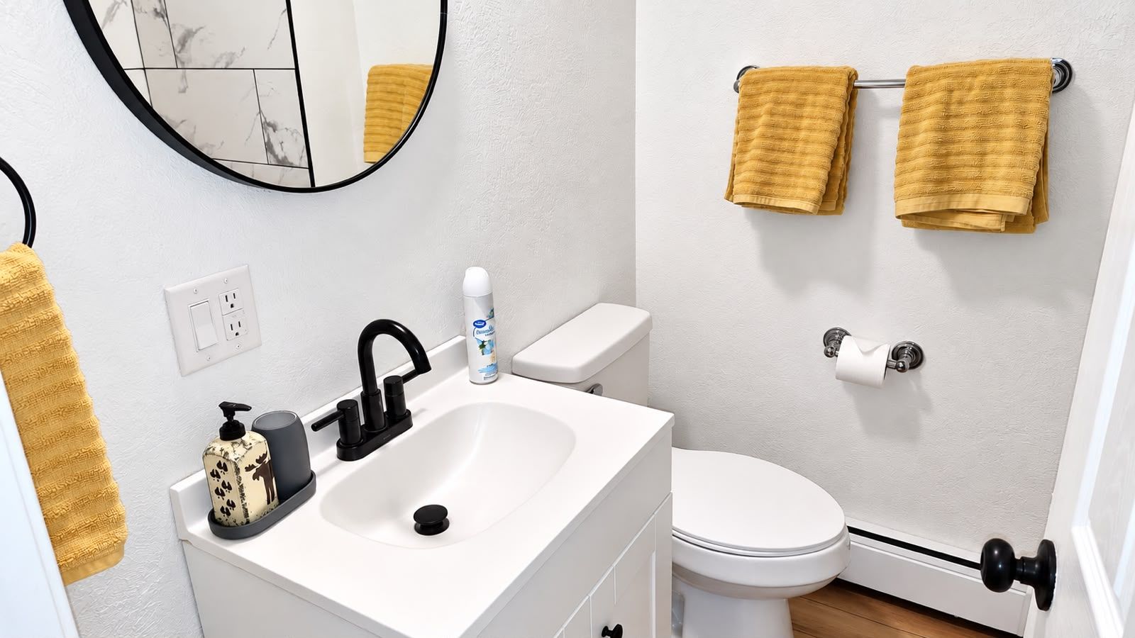 Un baño con lavamanos blanco, grifo negro y un inodoro. Hay dos toallas colgadas en la pared.
