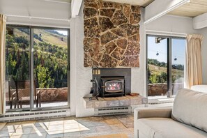 Ein Wohnzimmer mit Kamin aus Stein, einer Couch und großen Fenstern mit Blick auf einen bewaldeten Hügel.