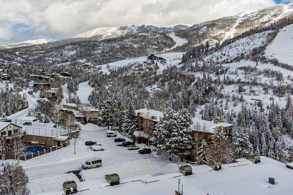 A snow-covered mountain resort with multiple buildings, a ski lift, and a parking area.