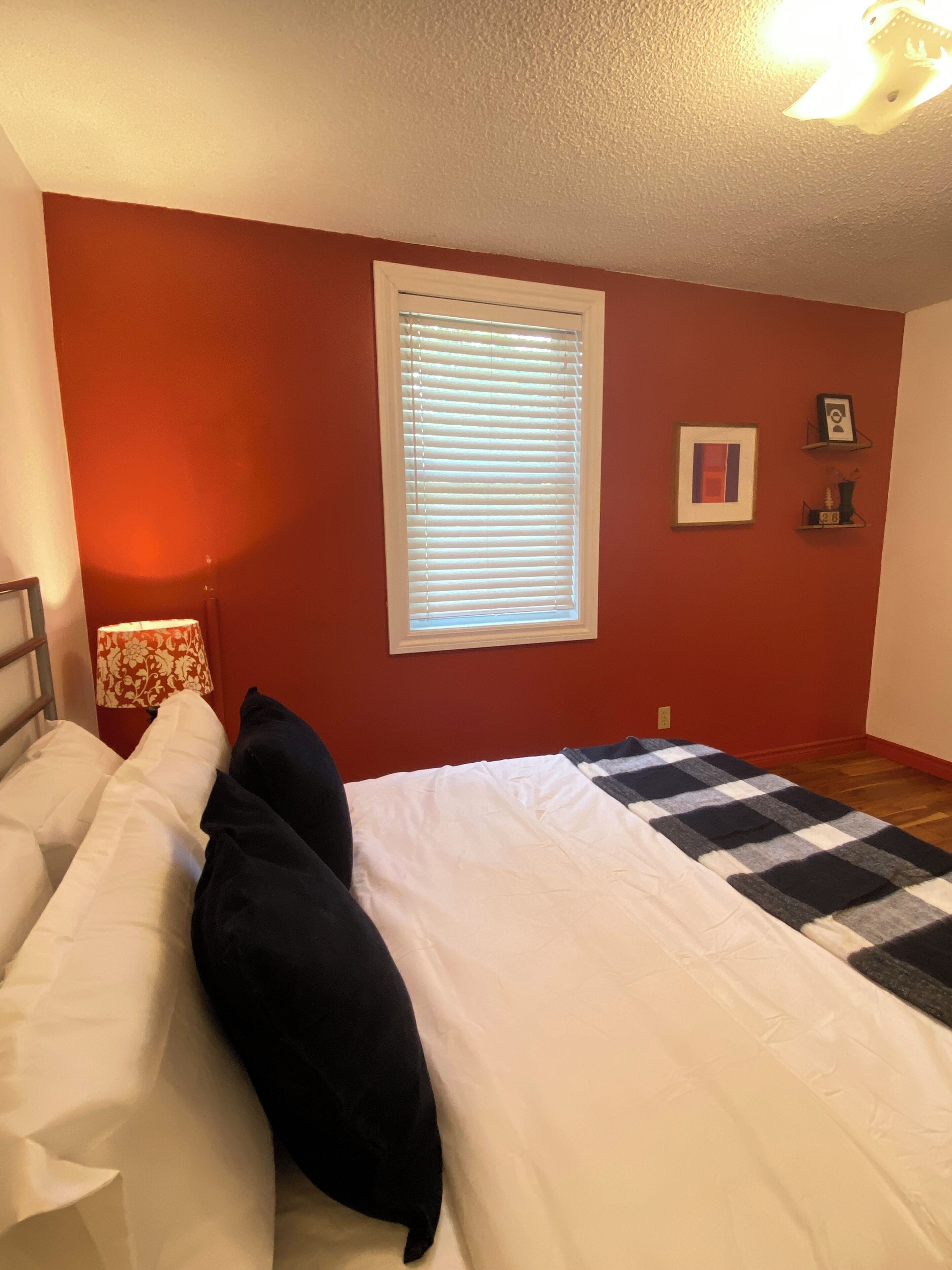 A bedroom with a bed, pillows, a window with blinds, a lamp, and a framed picture on the wall.