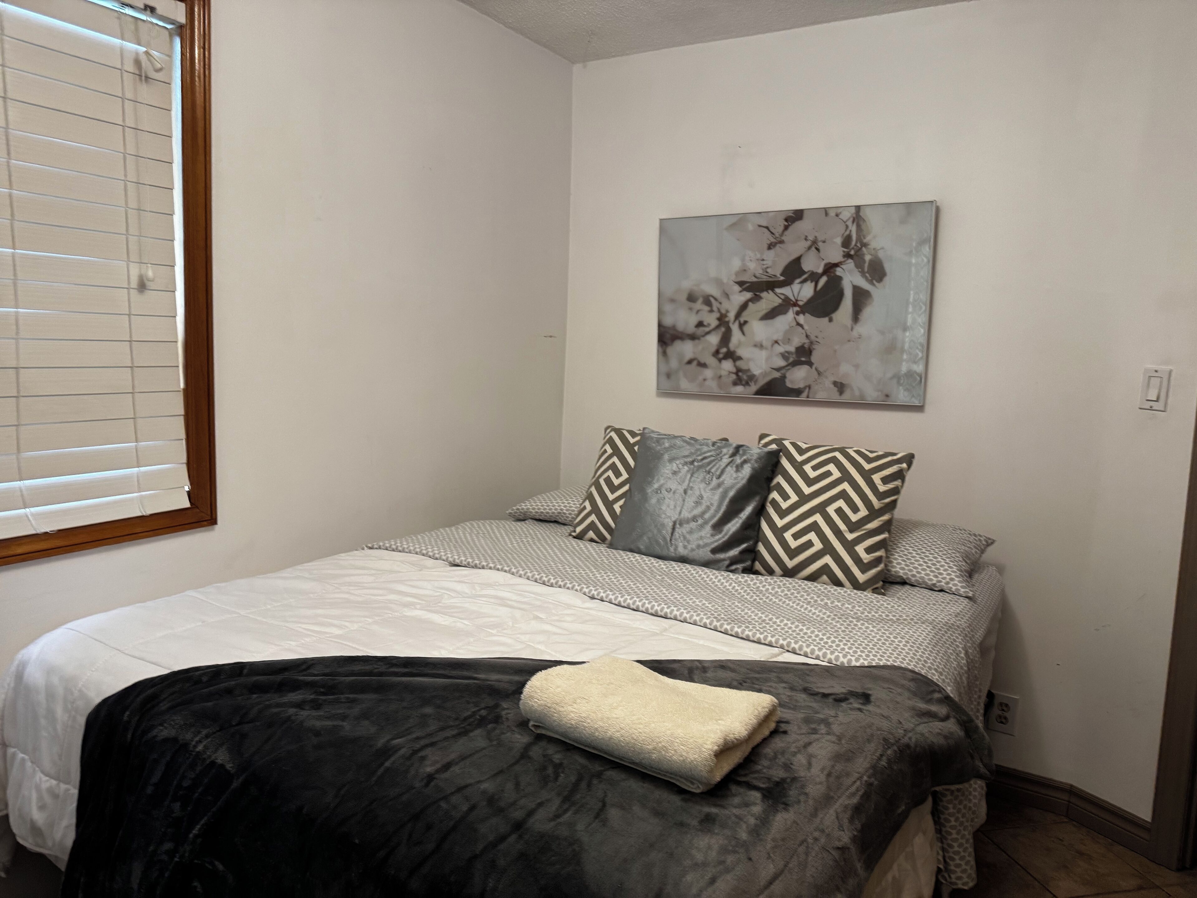 Ein Schlafzimmer mit Bett, Fenster mit Jalousien, einem Bild an der Wand und einer Decke auf dem Bett.