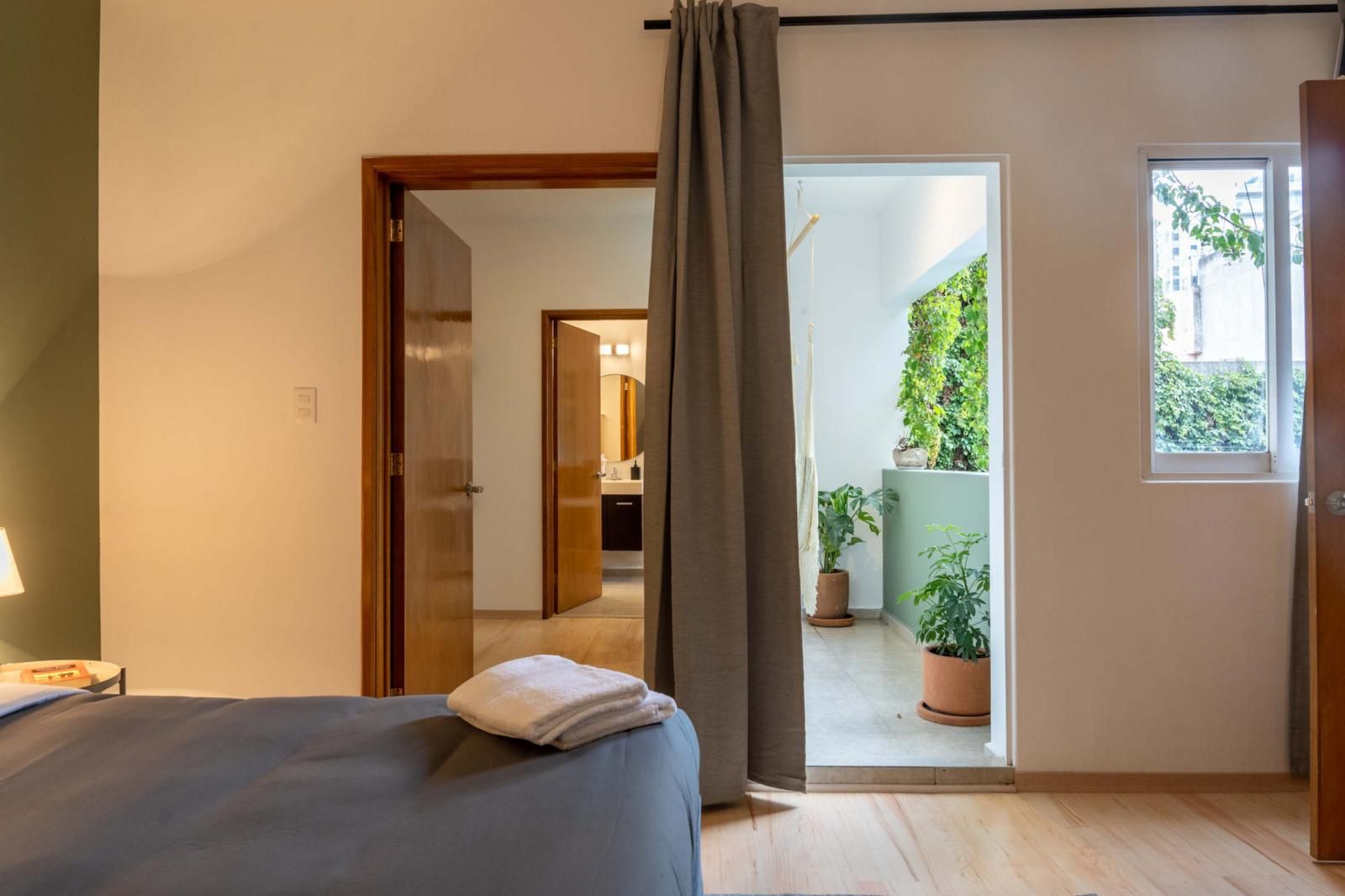 Un dormitori amb un llit, una nitzeta, una finestra i vista a un balcó amb plantes en testos.