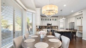 A modern kitchen with white cabinetry, a central island, and a dining area with a round table and chairs. - Golf Course Oasis | Pool, Spa, EV & Family Fun (Goodyear)