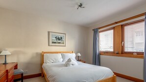 Un lit bien fait, avec du linge de lit blanc, une tête de lit en bois et une table de chevet sur laquelle se trouve une lampe. Un tableau encadré est accroché au mur au-dessus du lit.