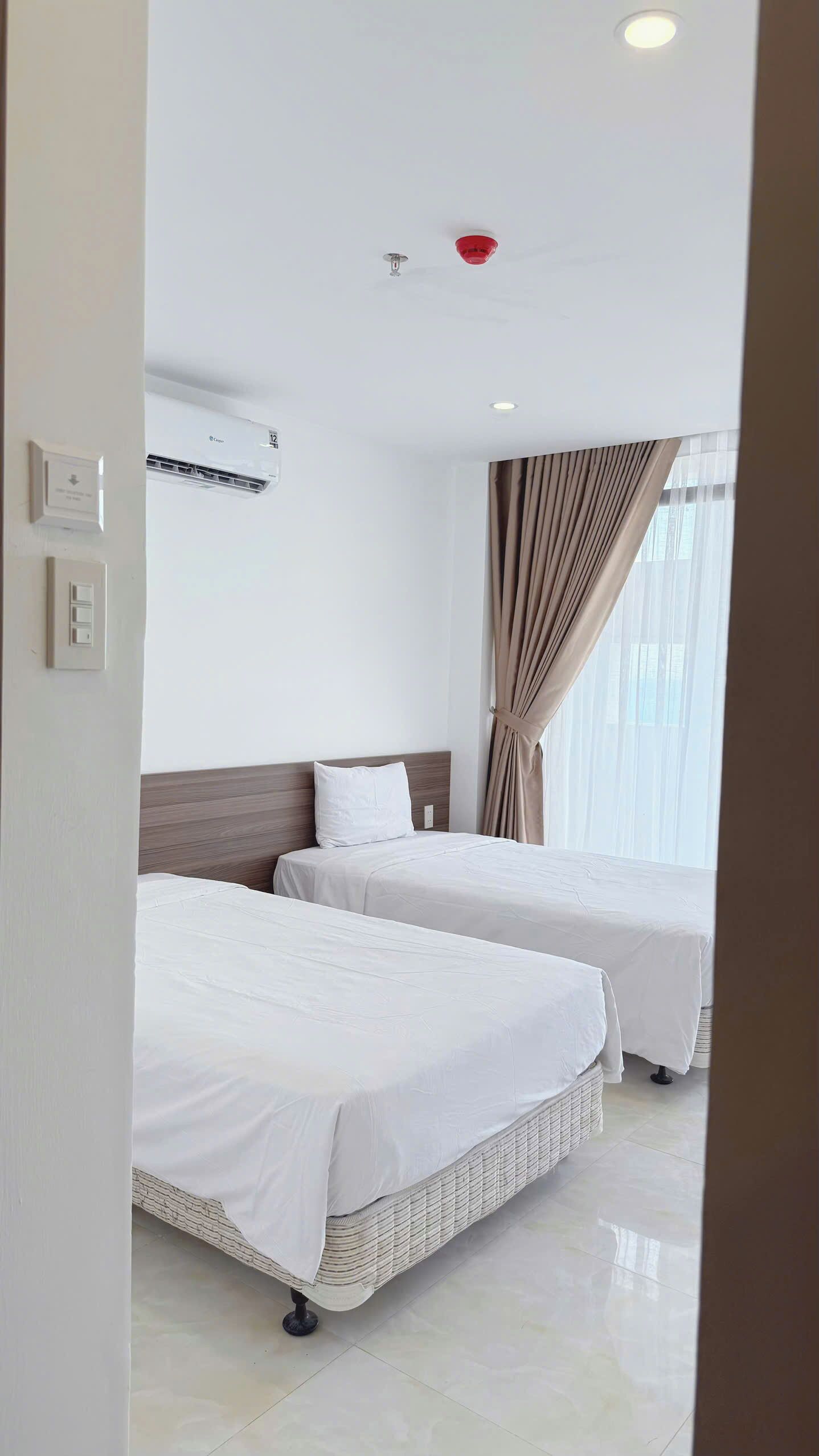 A hotel room with two beds, a ceiling light, an air conditioning unit, and a window with curtains.