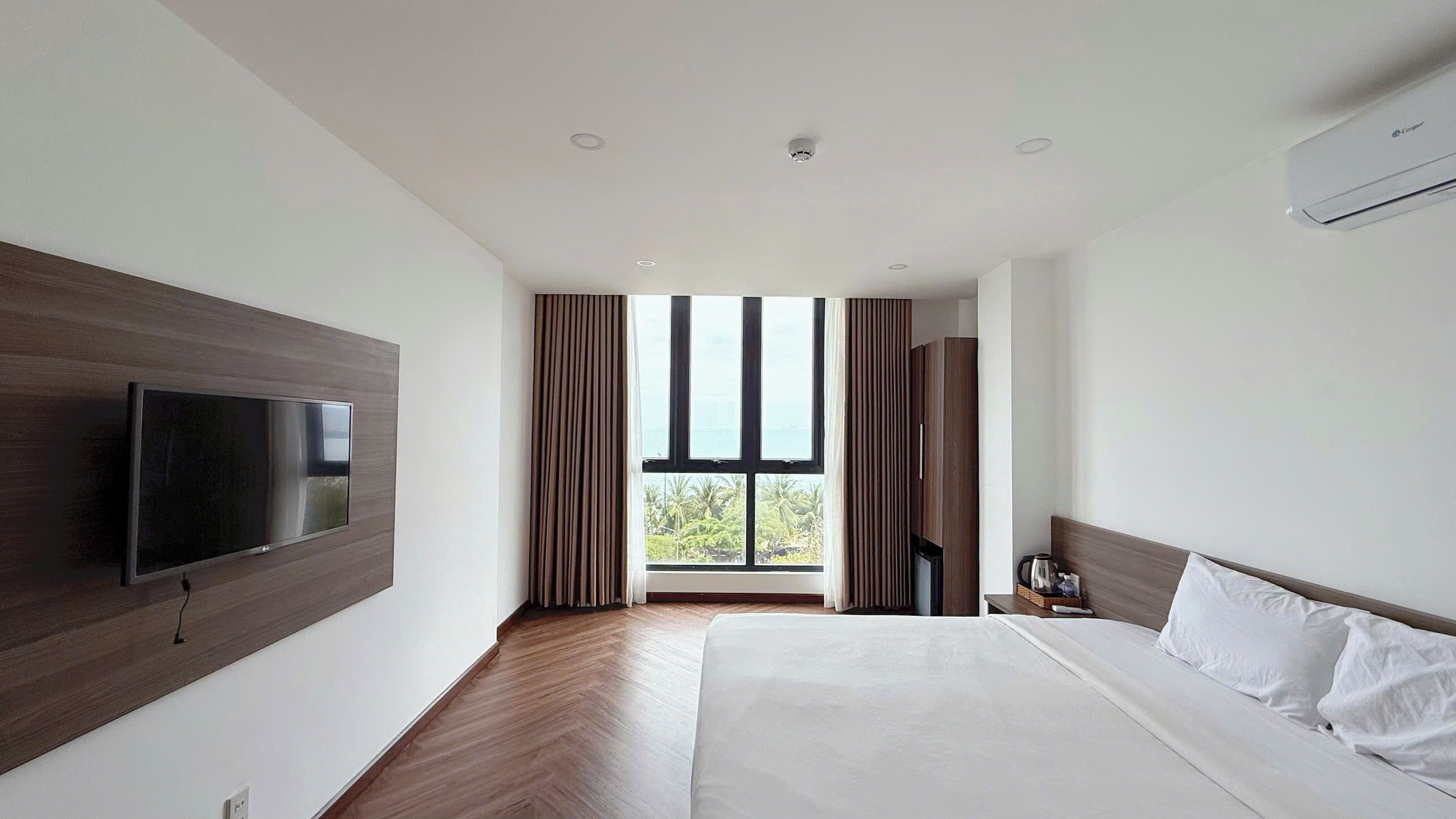 A modern hotel room with a large bed, a flat-screen TV mounted on the wall, a window with curtains, and an air conditioning unit.