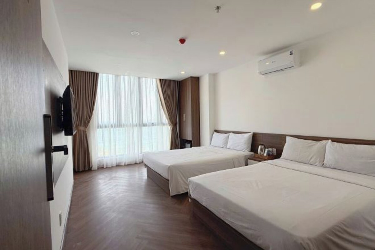 A hotel room with two beds, a flat-screen TV, an air conditioning unit, and a window with curtains.