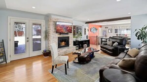 A spacious living room with a fireplace, leather sofas, and a large TV mounted above it. - DU Haven- Hot Tub, Outdoor TV Lounge &3 Full Baths (Denver)