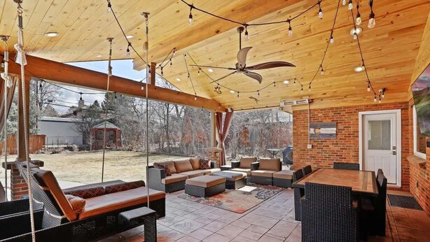 An outdoor patio with a wooden ceiling, ceiling fan, and string lights. There are multiple seating areas including sofas and a dining table. - DU Haven- Hot Tub, Outdoor TV Lounge &3 Full Baths (Denver)
