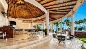 Un espacio al aire libre techado con mesas y sillas, una recepción y una piscina con palmeras y flores al fondo.