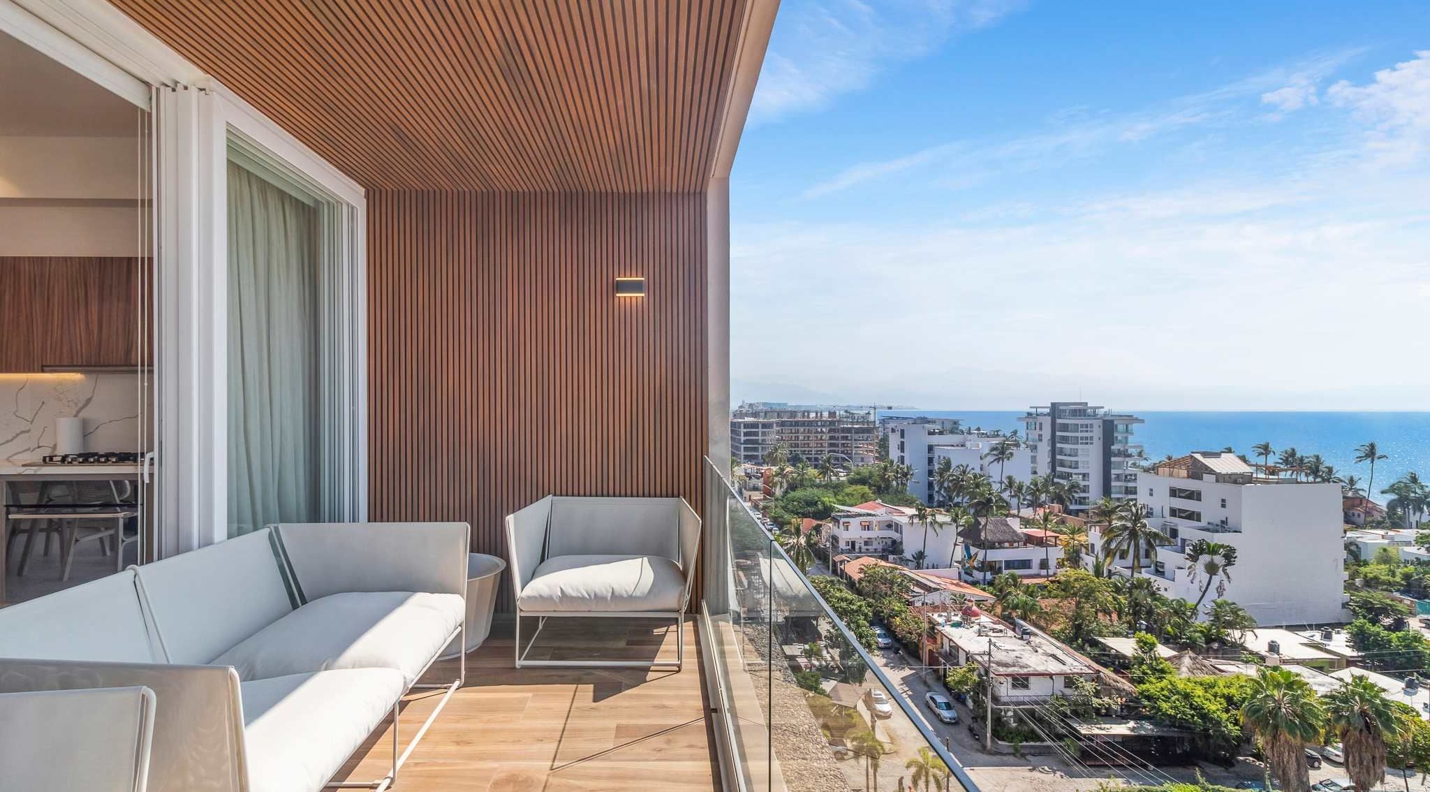 Un balcon doté d’un plafond en bois, d  ’un canapé et d’un fauteuil, offrant une vue sur un paysage urbain côtier avec des bâtiments et des palmiers.
