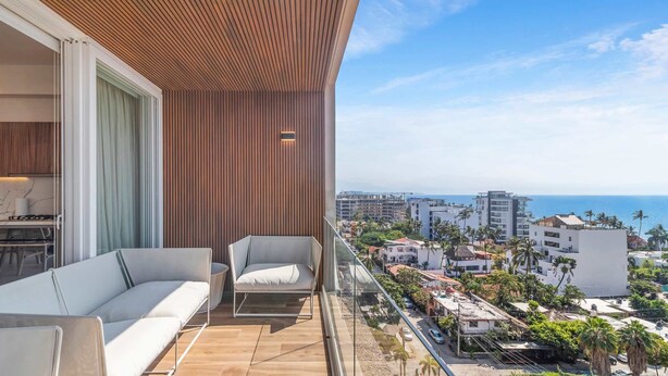 A balcony with a wooden ceiling, a sofa, and a chair, overlooking a coastal cityscape with buildings and palm trees. - Maralma 903: Panoramic Views and Rooftop Pool (Bucerías)