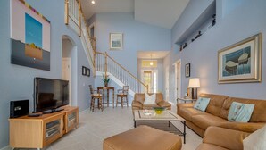 A spacious living room with a large TV, a sofa, a coffee table, and a staircase. - Highly Desired Gated Community! Close to Beaches! Private Heated Pool w/ Golf Course Views! (Fort Myers)