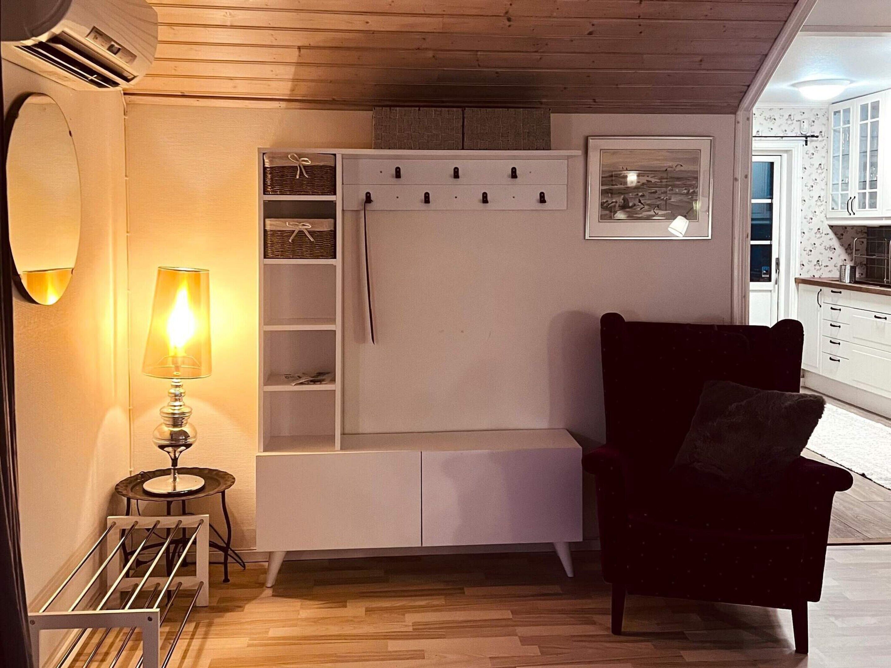 Un salón acogedor con un sillón oscuro, un mueble blanco para entretenimiento, una lámpara y una mesita con una copa de vino.