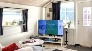 Un salon confortable avec un téléviseur à écran plat, un canapé blanc, une table basse et une petite table d’appoint. Des plantes en pot sont posées sur le rebord de la fenêtre, et une porte avec un meuble à chaussures se trouve à côté.
