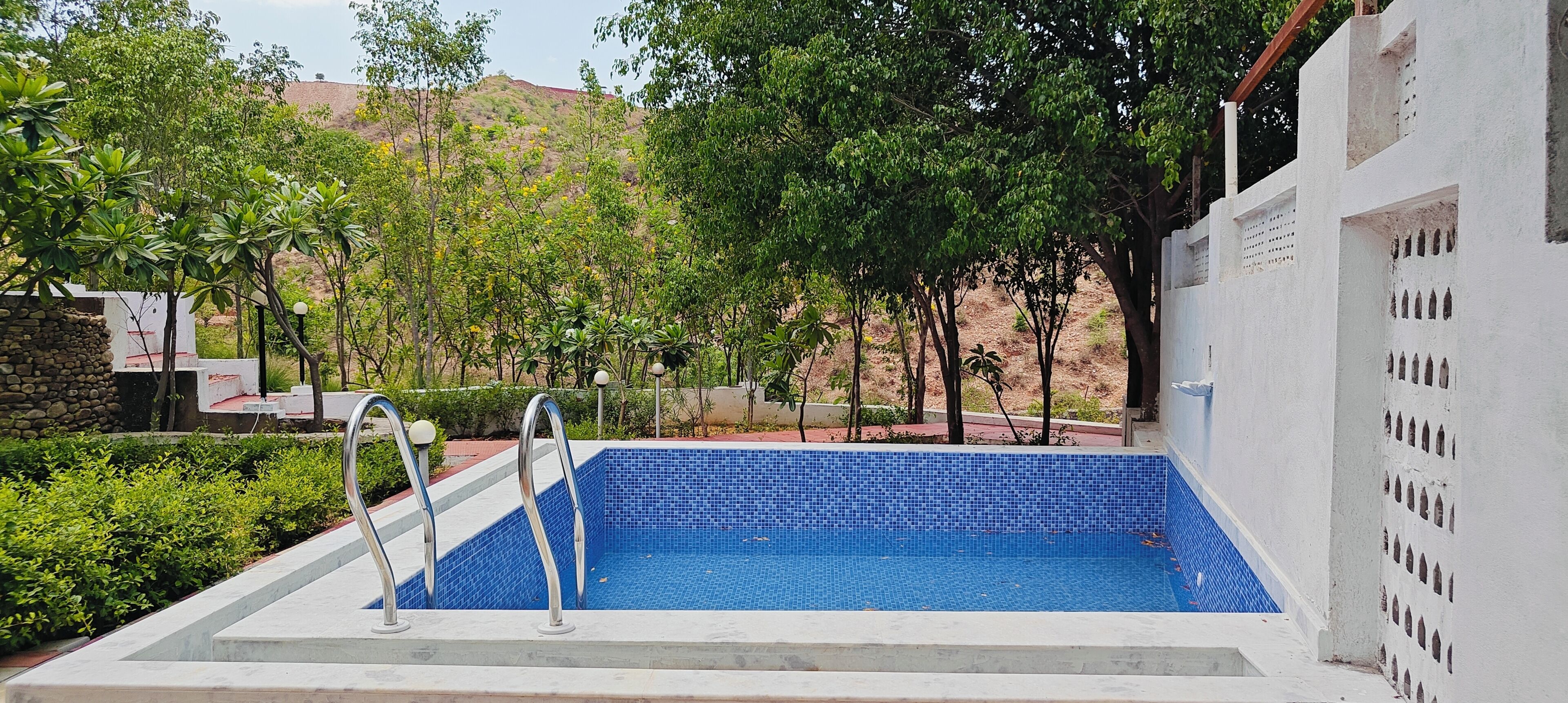 A swimming pool with metal ladders, surrounded by greenery and a white building with decorative cutouts.