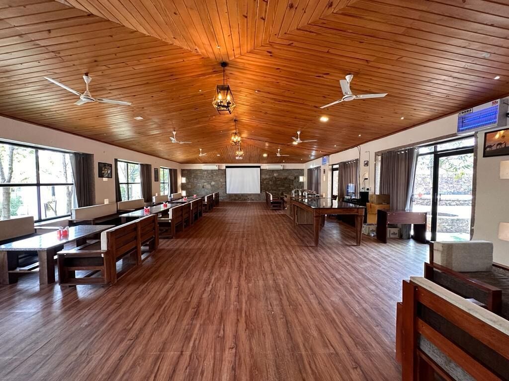 A spacious indoor area with wooden floors and ceiling fans, featuring long tables and benches, large windows, and a stone wall in the background.