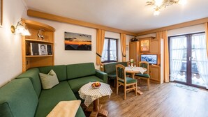 Een knus ingerichte kamer met een groene bank, een klein tafeltje met een tafelkleed met stippen, een houten kast en een schilderij van een zonsondergang.