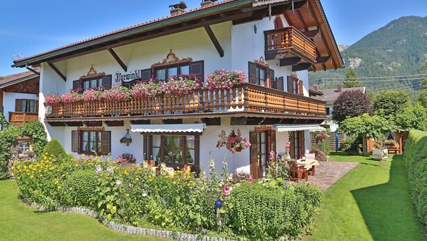Un chalet tradicional con balcón, un jardín lleno de flores y un paisaje de montañas al fondo.