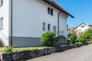 Un edificio de dos pisos con un muro de piedra en primer plano, cielo despejado y una casa vecina.