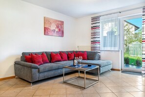 Un salón con un sofá gris, cojines rojos, una mesa de centro de cristal, un cuadro en la pared y vistas a través de una ventana.