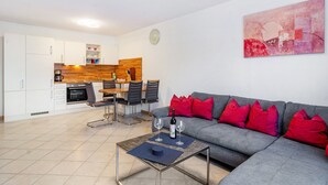 A modern living room with a grey sofa, red cushions, a dining area with a wooden table and chairs, and a kitchen in the background.