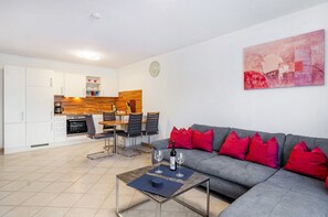 Un salon moderne avec un canapé gris, des coussins rouges, un coin repas avec une table et des chaises en bois, et une cuisine en arrière-plan.