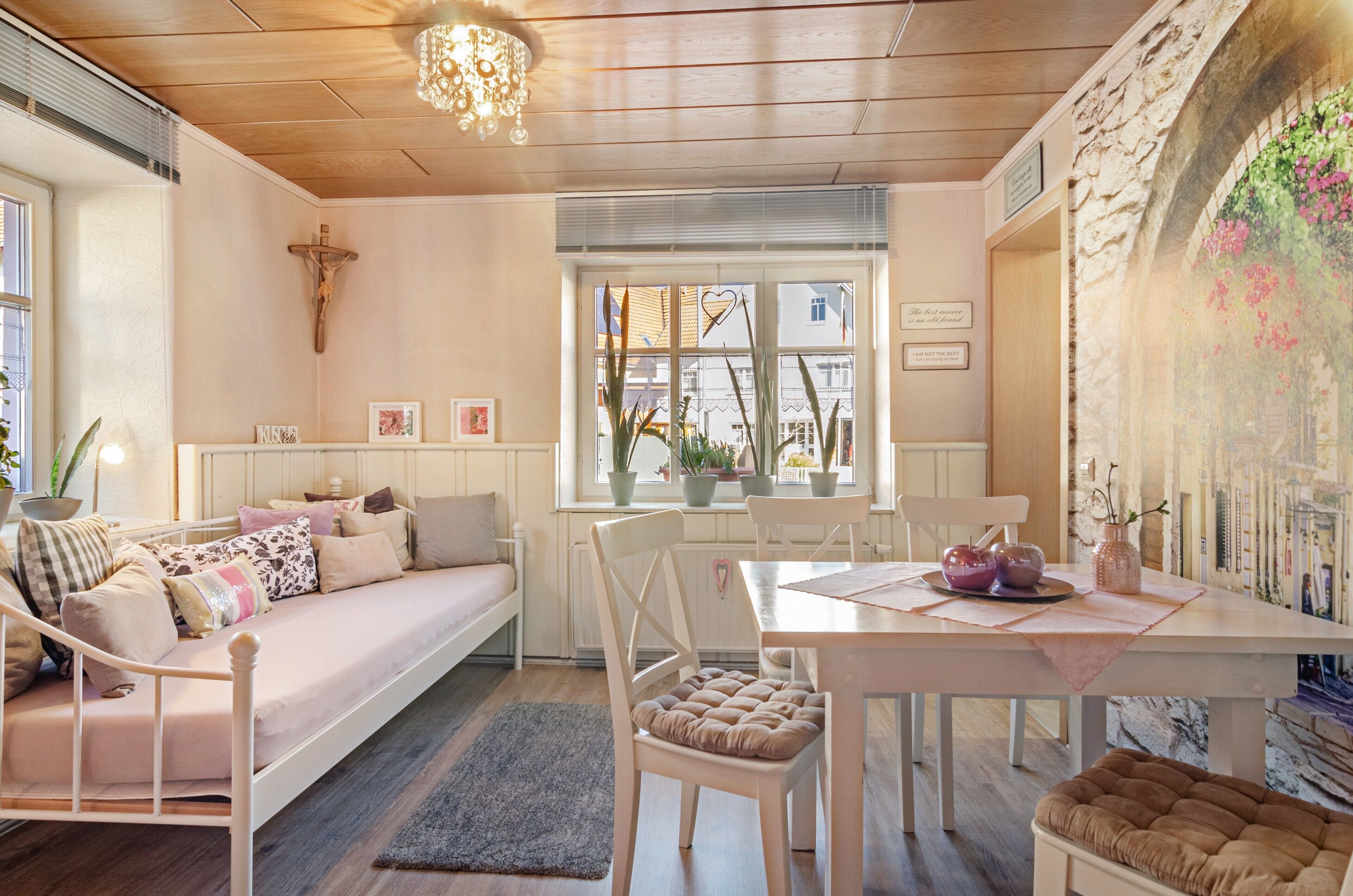 Una habitación acogedora con cama, una mesa de comedor, sillas, una araña y una ventana con plantas.