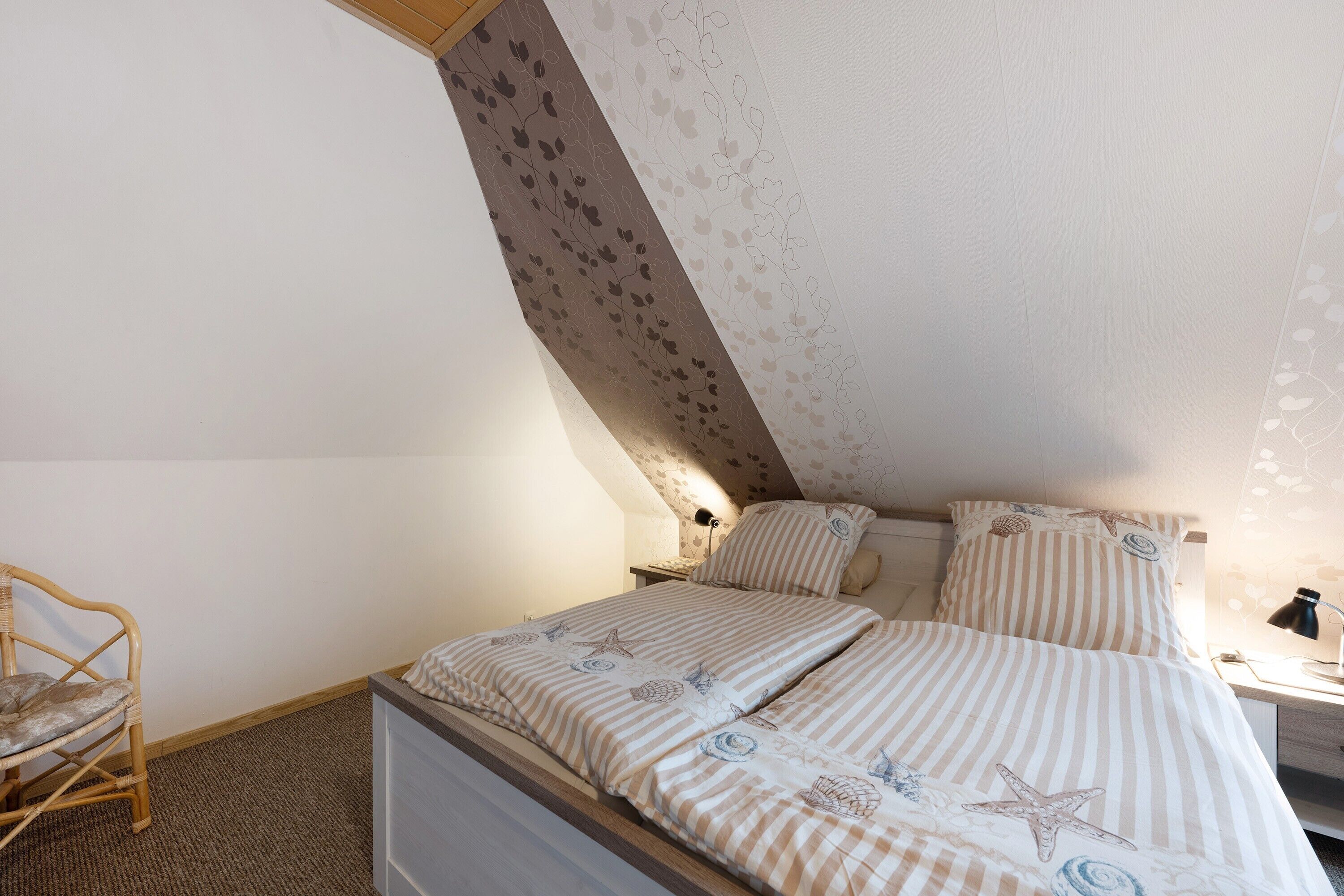 Een slaapkamer met een schuine kap, een bed met gestreepte beddengoed, een stoel en een nachtkastje met een lamp.