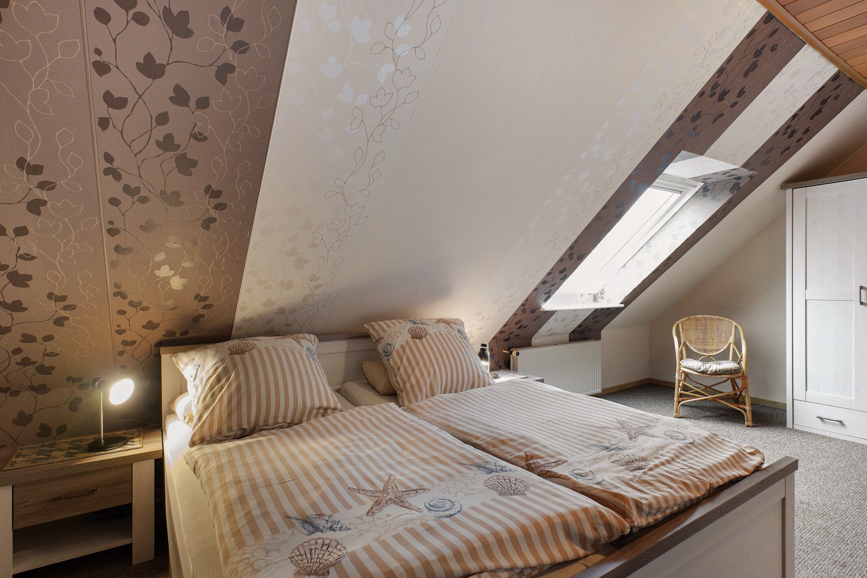Une chambre avec un plafond en pente, un lit avec une literie à rayures, une lampe de chevet, une chaise en osier et une armoire.