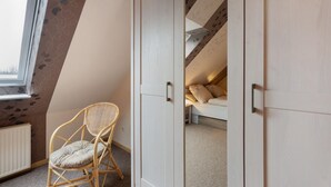 Ein Zimmer mit schrägem Dach, einem Korbstuhl, einem Kleiderschrank und einem Bett mit Kissen.