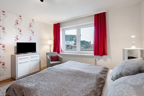 Une chambre à coucher avec un lit, un téléviseur posé sur une commode, une chaise, une fenêtre avec des rideaux et une lampe.