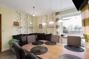 Un coin repas avec une table en bois, des sets de table noirs, un canapé et une fenêtre avec des rideaux.