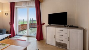 A room with a TV on a cabinet, a table, and a view through a glass door. - Apartment 'Deichblick 3' with Shared Garden and Balcony (Hedwigenkoog)