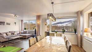 Een moderne woonkamer met een grote houten eettafel, een bank en zicht op een besneeerd landschap door grote ramen.