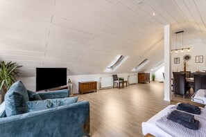 A spacious loft apartment with a high ceiling, skylights, a large TV, a sofa, a dining area, and a bed.