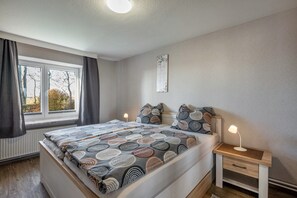 A bedroom with a bed, bedside tables, a window with curtains, a radiator, and a wall-mounted artwork.