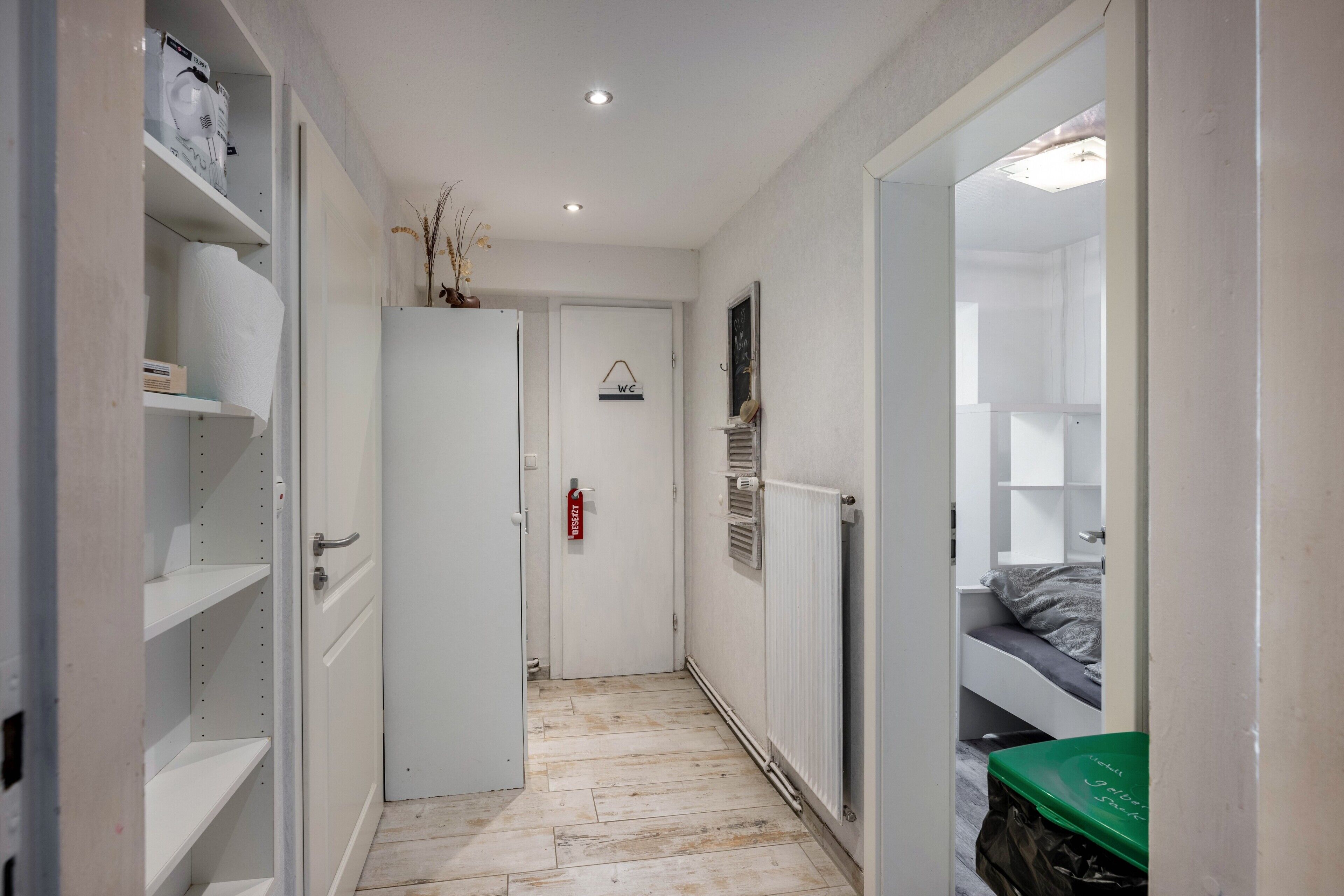 Un pasillo estrecho con una puerta blanca, un radiador y una cama con colchón gris en la habitación contigua.