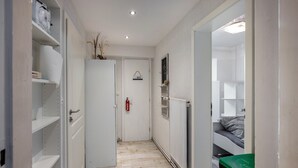 Un pasillo estrecho con una puerta blanca, un radiador y una cama con colchón gris en la habitación contigua.