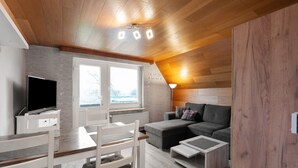 Un salon moderne avec un plafond en bois, un canapé gris, une table à manger blanche avec des chaises et un téléviseur à écran plat.