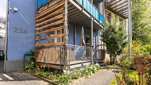 A building with a blue facade, a balcony with wooden scaffolding, and a garden area with flowers and plants.