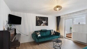 En moderne stue med sofa, sofabord, et fladskærms-tv monteret på væggen, et klaver og et stort vindue.