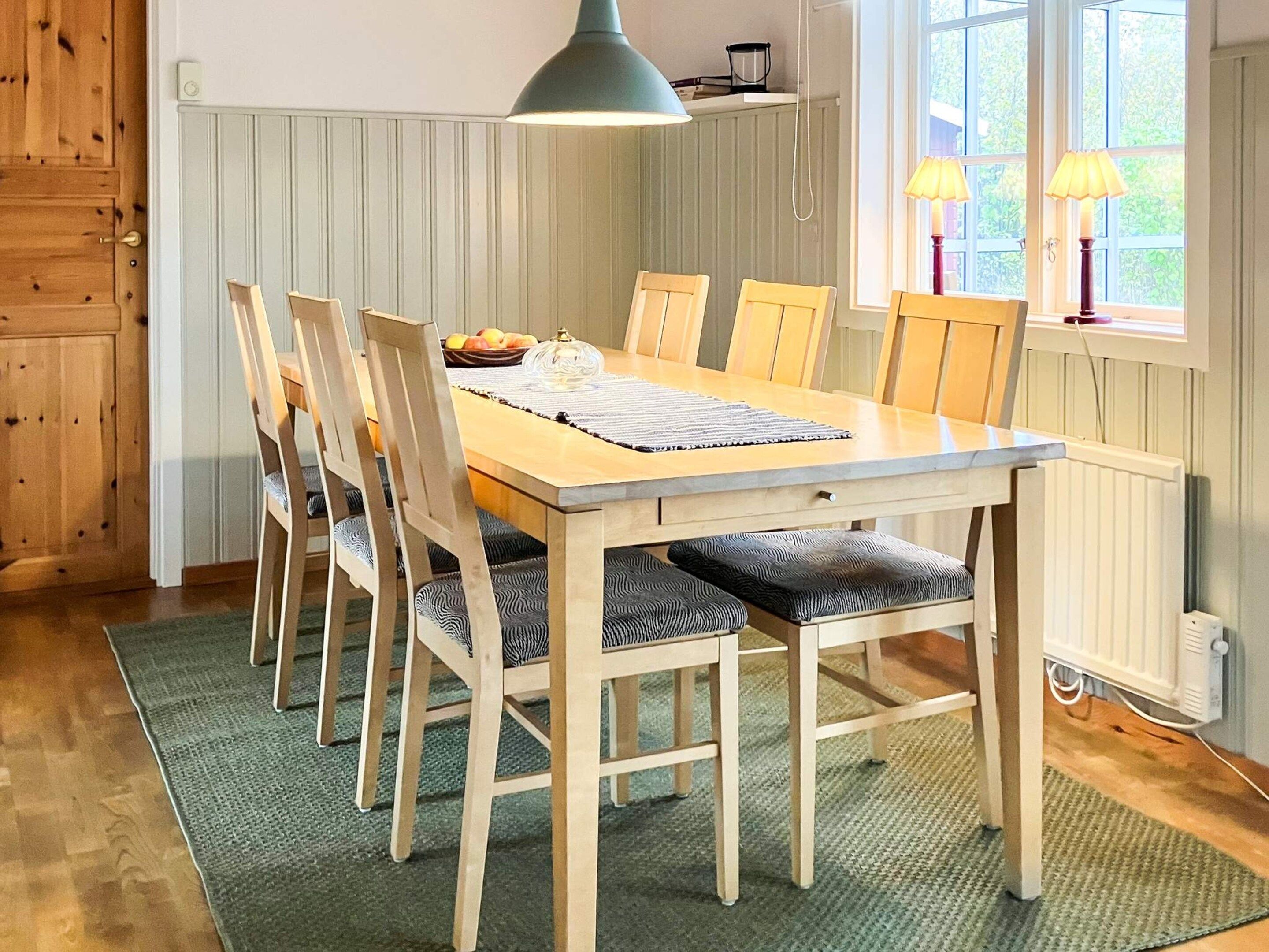 A dining area with a wooden table and chairs, a green rug, and a window with a view of greenery.